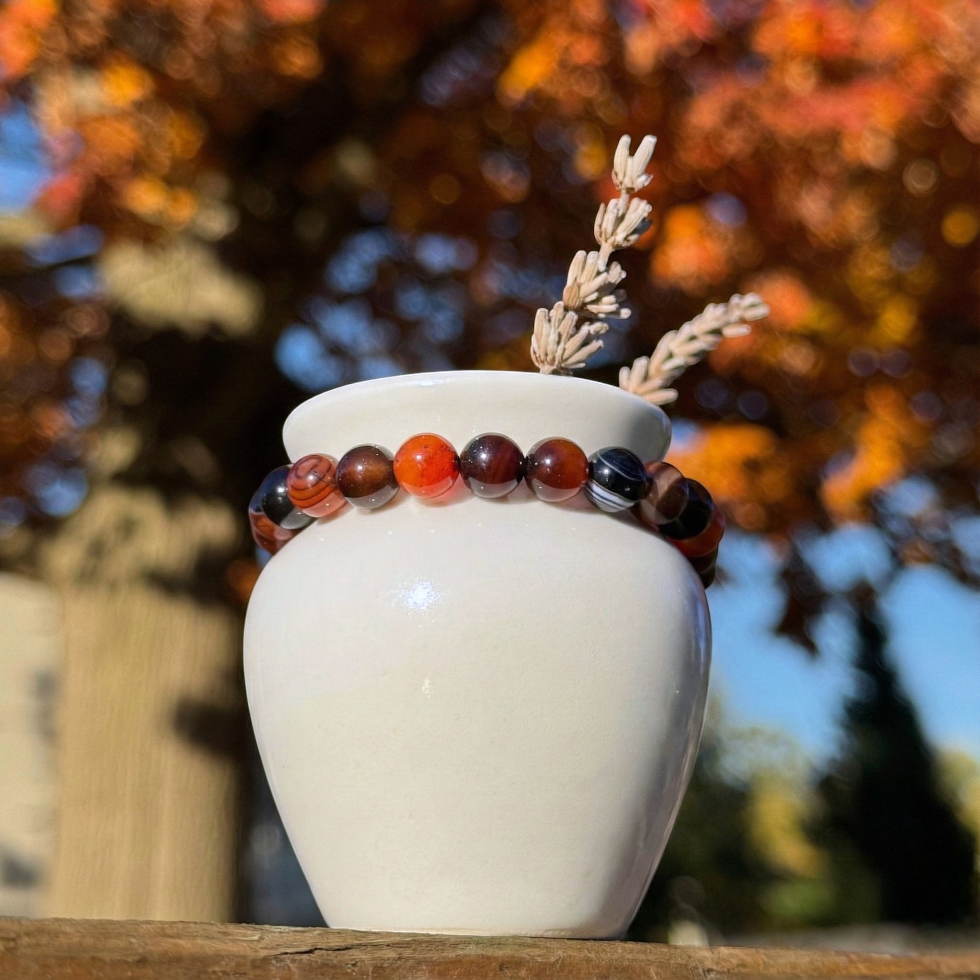White vase with a red banded agate bracelet and dried plants against an autumn tree background